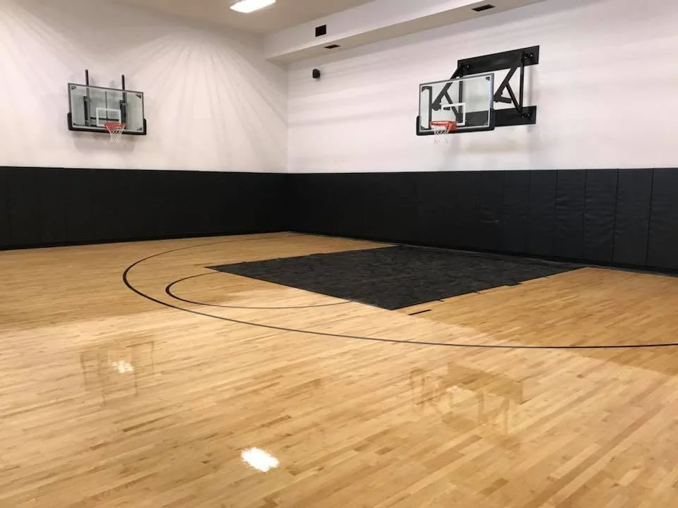 Indoor basketball court with polished wood flooring, black wall padding and two hoops