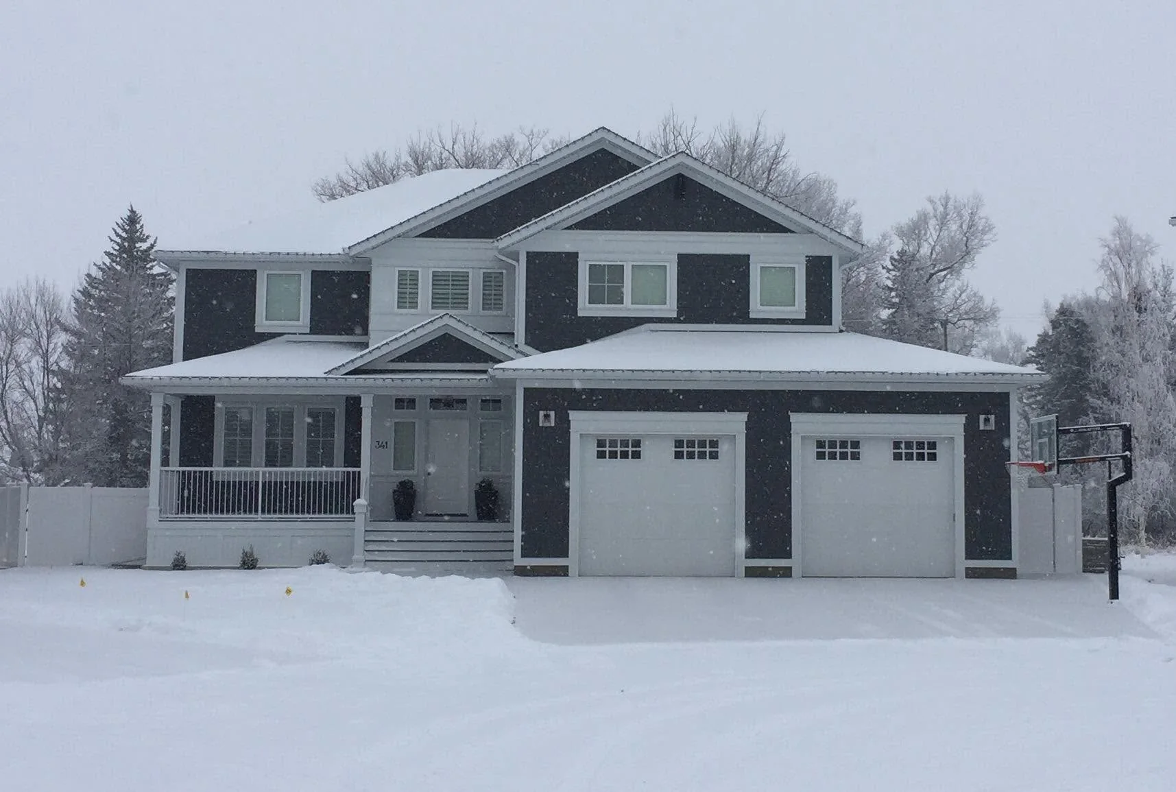 Dark two-storey home with a covered porch and two garage doors during snowfall