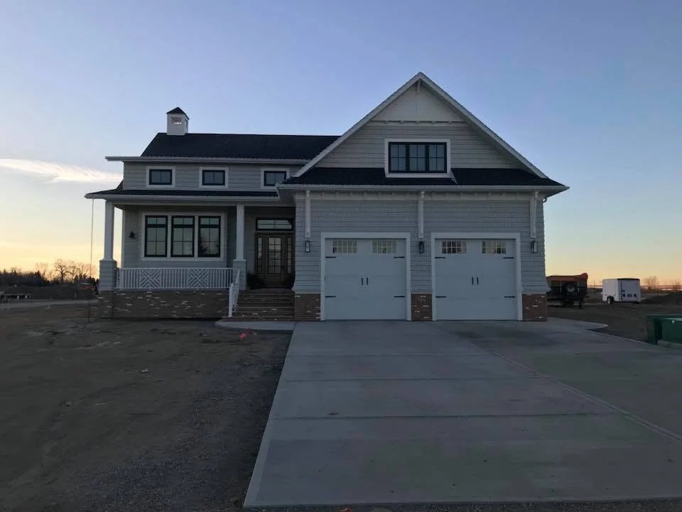Shingled home at dusk with a covered porch, brick base and two garage doors
