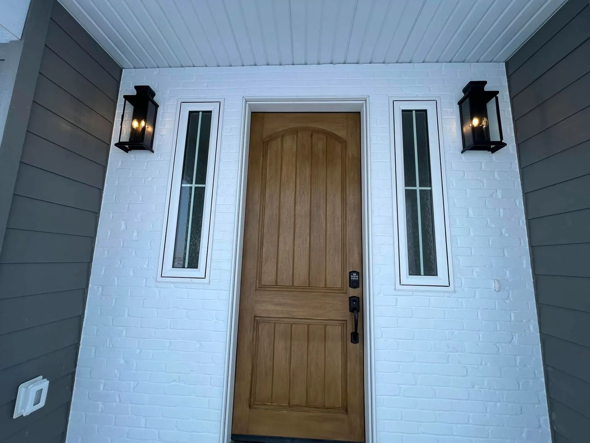 Wood front door between narrow sidelights, with black lantern sconces on white brick